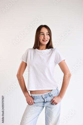 Confident young woman in white t shirt and light jeans standing against clean white background with hands on hips, relaxed smile, casual fashion pose, bright studio portrait with copy space