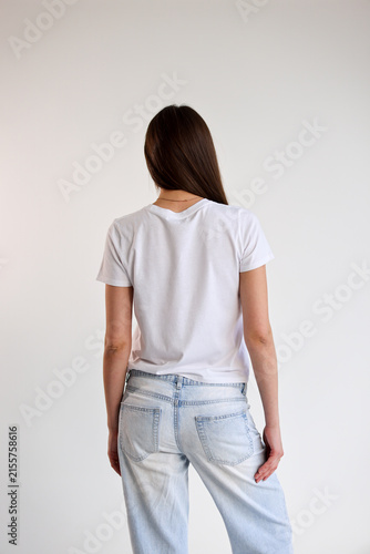 Rear view young woman standing against clean white backdrop wearing plain white t shirt and light washed jeans with relaxed posture minimal studio composition and generous copy space for fashion and