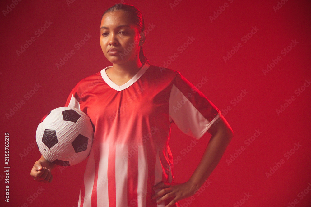 Obraz premium Adult African woman standing in red studio wearing red-white jersey holding black-white ball