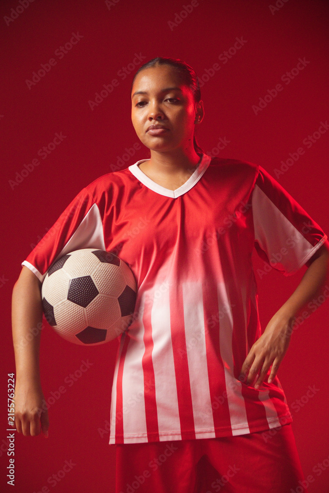 Obraz premium African American woman posing in studio wearing red-white kit, holding black-white ball at hip