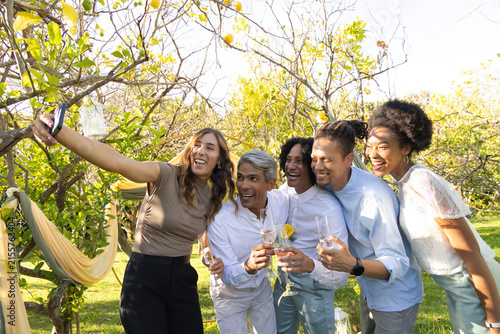 Group of friends taking selfie in garden, holding smartphone and champagne flutes under lanterns
