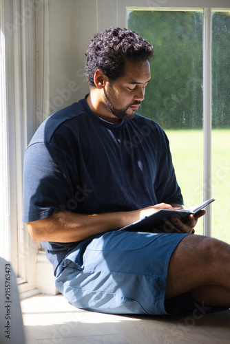 Mid adult African American man sitting on window seat at home reading book with sunlight streaming