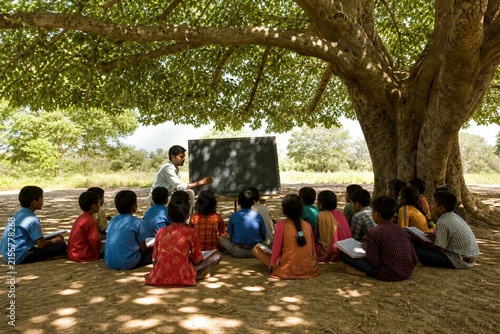 Indian male teacher conducting an outdoor lesson for schoolchildren under a large tree.