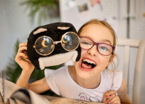 Playful girl with glasses laughs with a plush toy wearing funny spectacles at home. Happy child poses beside an open book, creating a joyful, humorous childhood and reading scene.