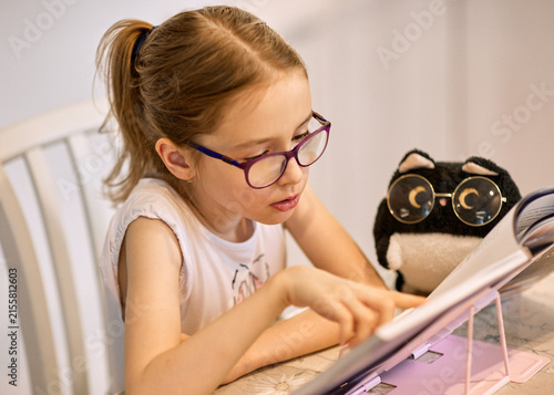 Focused girl with glasses enjoys reading a book at home, pointing at the page beside a plush toy. Young child in a white shirt practices learning and literacy in a cozy indoor setting.