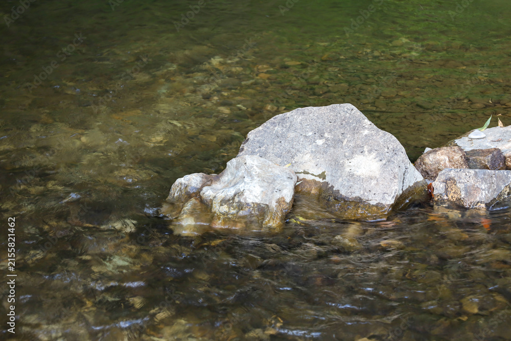 Naklejka premium A large gray stone emerges from the transparent water of a mountain or forest river. The clear current gently washes over smooth stones and small pebbles at the bottom. Natural lighting creates a sens