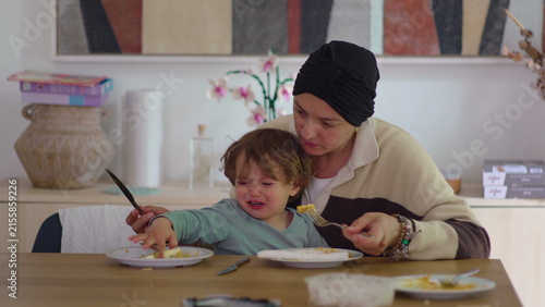 Fotografía Mother offering food to reluctant toddler at dining table in family home during