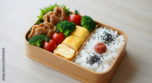 Bento box filled with rice, tamagoyaki, and vegetables sits on a light wooden table.