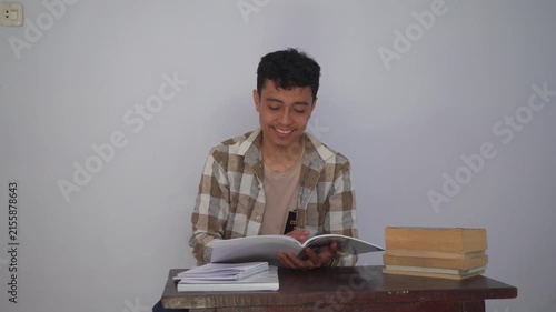 Cheerful Young Man Sitting at a Desk Reading a Book with a Stack of Textbooks and Papers