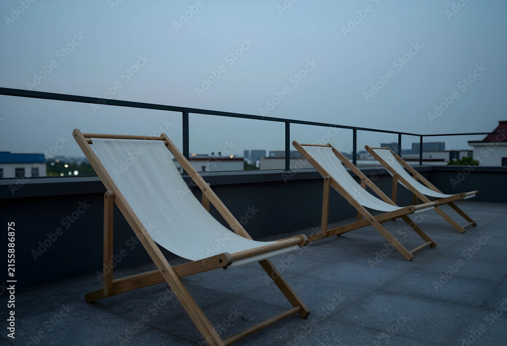 Fototapeta premium Three empty wooden deck chairs with white canvas on a rooftop terrace at dusk, urban skyline in the background, the urban skyline with buildings and a dark sky forms the background