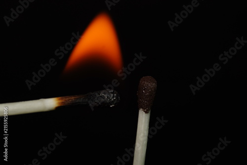 Conceptual macro photograph of two matches meeting after combustion, with a glowing flame in the background and dramatic black negative space.
