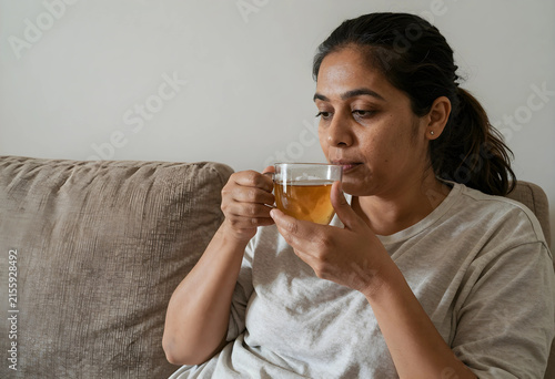 Indian woman drinking hot tea from a clear glass mug while relaxing on a comfortable sofa at home