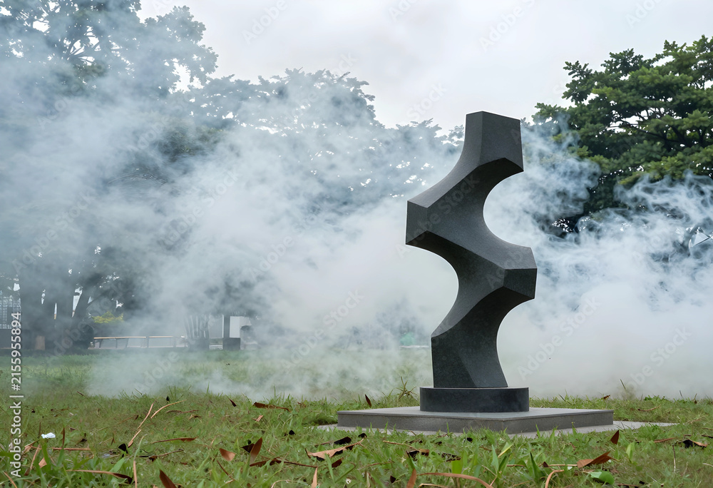 Naklejka premium Dark gray stone sculpture with geometric shapes in a park with Smoke and trees, Smoke drifts around the artwork and trees in the background