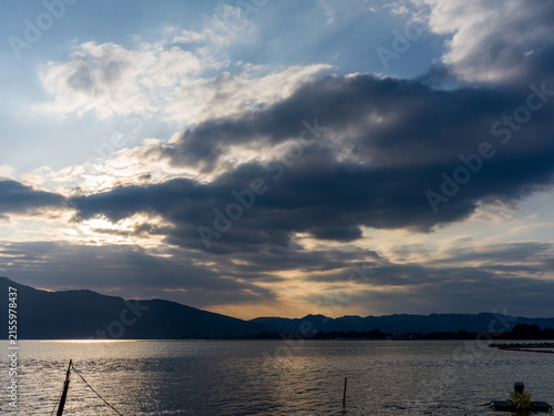 Quadro su tela Wide lake landscape under dramatic cloudy sky catching golden sunset rim