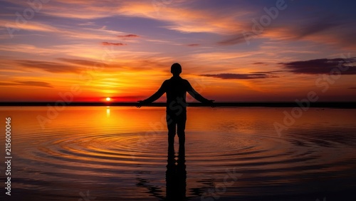 Silhouette of person standing in calm water at sunset.