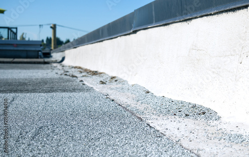 Flat roof parapet with granule loss on modified bitumen cap sheet. Low angle view. Aggregate pile visible at base. SBS 2-ply roofing system requiring maintenance. Selective focus.