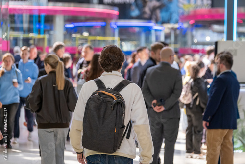 Fotografía Backpack-wearing visitor walks into a crowded exhibition hall filled with people and colorful illuminated booths
