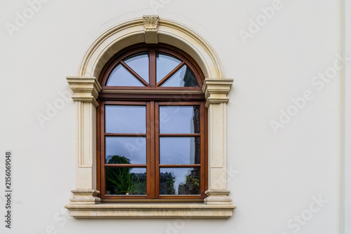 Fotografía A classic architectural window with a rich brown wooden frame, a rectangular grid section paired with an adjacent arched fanlight, all set within an ornate, stepped beige masonry, stucco surround