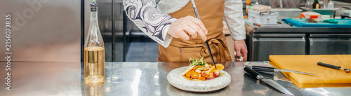 Chef in a professional kitchen plating a gourmet dish with colorful vegetables on a white plate, stainless steel countertop and kitchen tools visible in the background