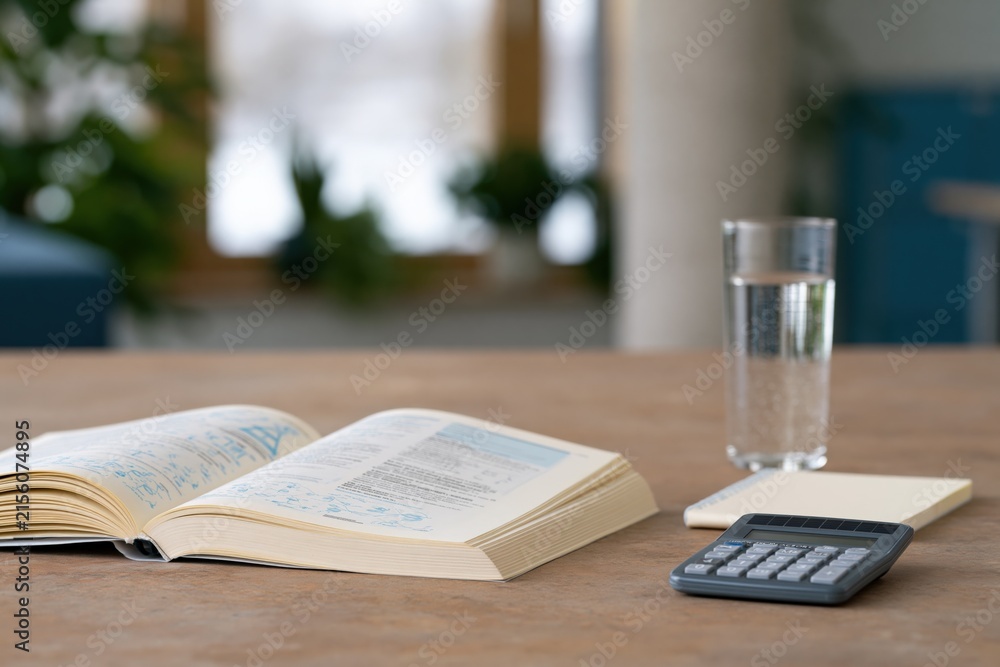 Obraz premium Open textbook and calculator on wooden table with glass of water for study workspace background and finance poster design