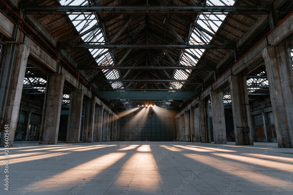Fototapeta premium Vast abandoned factory hall with sunbeams through high windows and long floor shadows for wallpaper and banner background