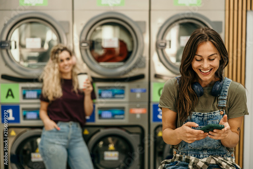 Happy women friends using smartphone at self-service laundromat