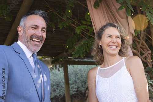 Couple laughing under wooden pergola in garden, light-blue suit and white-lace dress, lens flare