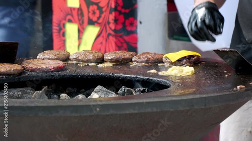 Grilled burgers sizzle over glowing charcoal. Chef's gloved hand places cheese slices. Fatty patties cook in open flame. Food vendor prepares quick meal. Perfect for food festival or street market ads