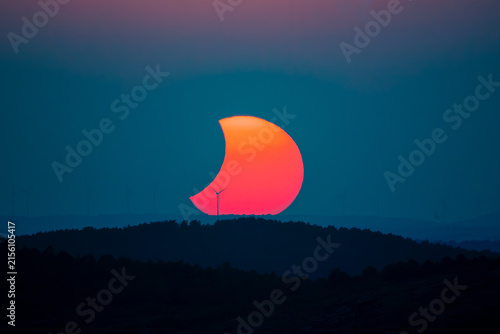 Partially eclipsed sun setting behind wind turbines and forested hills at dusk