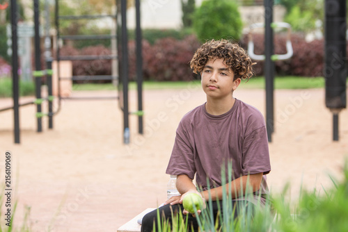 Teen athlete resting after workout while holding a green apple as a healthy snack in a park. Thoughtful teenage boy taking a break after an outdoor workout and holding a fresh green apple