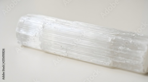 Long translucent selenite crystal wand displaying fibrous textures and bright white highlights under soft studio lighting on a neutral surface