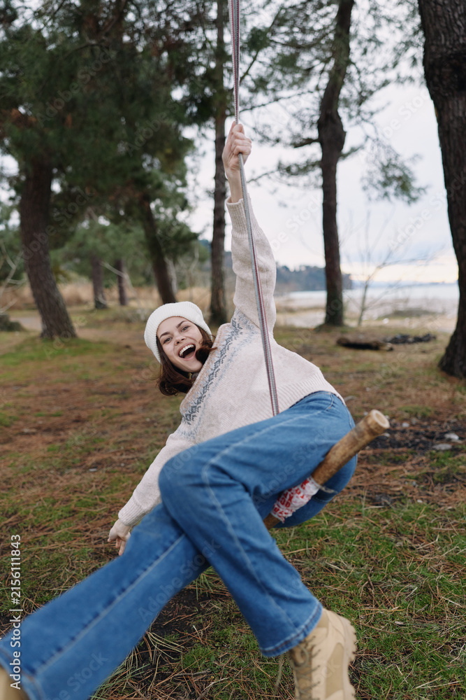 Obraz premium Joyful woman swinging outdoors in a forest park, laughing and leaning back on a rope seat, wearing a beanie, sweater and jeans. Vivid trees and grass create a carefree, candid moment of play.