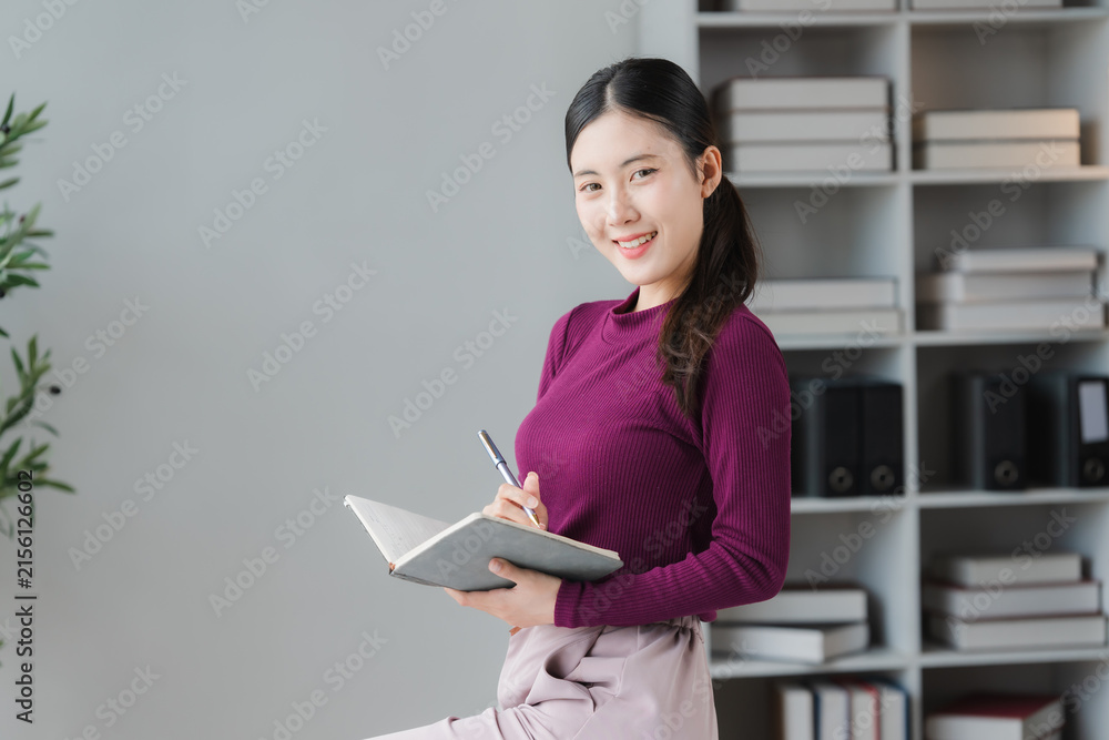 Fototapeta premium A Young Professional Woman Taking Notes: A cheerful young woman with dark hair pulled back smiles at the camera while writing in a notebook.