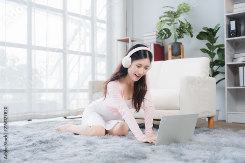 фотографія Young Woman Enjoying Music and Laptop: A smiling young woman wearing headphones sits on a plush carpet in a bright, modern living room, engrossed in her laptop and listening to music