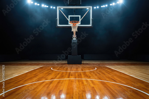 Empty professional basketball hoop and backboard over polished wooden court floor under bright arena stadium lights prepared for competitive tournament match play.