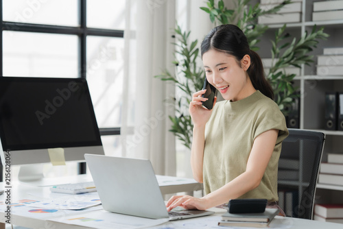 Fotografía Focused Professional: A young Asian woman with a cheerful expression, engaged in a phone call while working on her laptop at a modern office desk