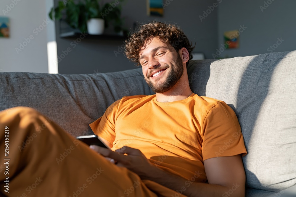Fototapeta premium Smiling man relaxing on sofa with smartphone in bright living room for lifestyle background and wellness banner design