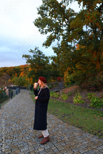 Young woman enjoying coffee while walking outdoors on a cobblestone path surrounded by colorful autumn trees.