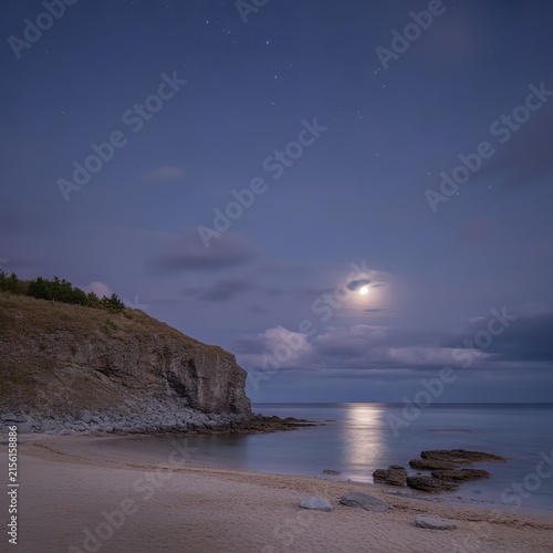 Full moon rising over a rocky coastal beach at night with calm ocean water and starry sky