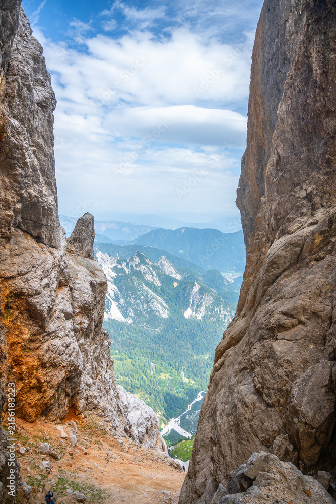 Fototapeta premium The scene shows a gap between two large rock formations in the Julian Alps. The view reveals green valleys and distant mountains under a cloudy sky. A person can be seen in the distance.