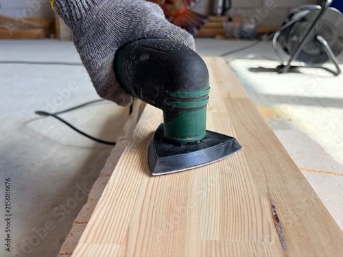 a worker's hand wearing a grey knitted protective glove operating an electric detail sander. Renovation or construction work