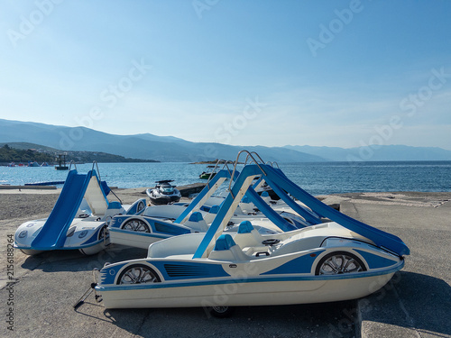 Pedal boats with blue canopies parked on a sunny beach by the sea, mountains in the background
