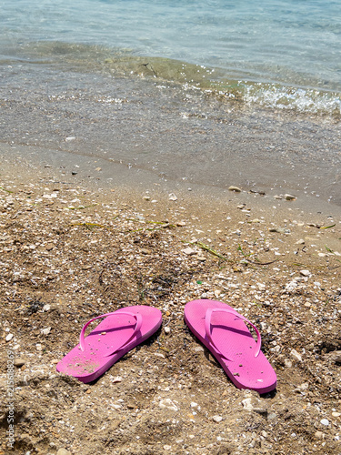 Pink flip flops on sandy beach shore with gentle waves and clear blue water in background