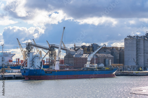 Large blue industrial cargo vessel moored at a harbor quay with deck cranes, loaded containers, and grain silos under a vast cloudy blue sky on a sunny day