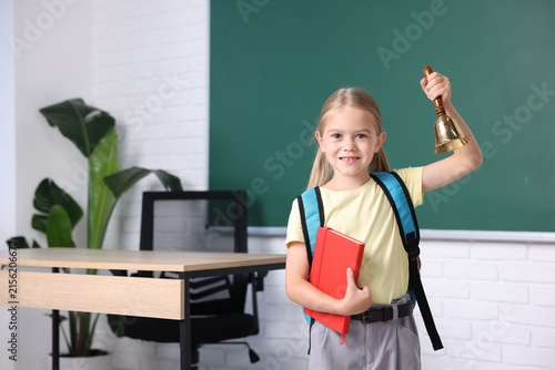Cute little girl with notebook ringing school bell in classroom. Space for text