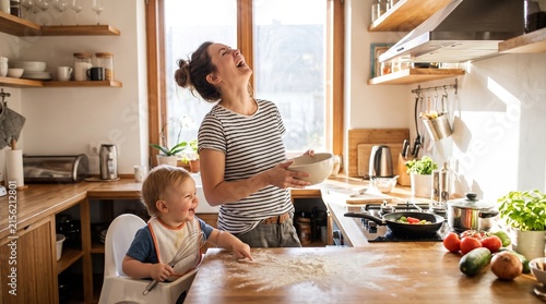 A mother and her baby boy laughing while making a mess in the kitchen, flour scattered on the table. Sunlight streams in from the window, illuminating the joyful scene