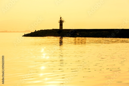 Lighthouse on a small rocky breakwater island silhouetted against a golden sunset sky and calm water, serene seascape, Mediterranean coast.
