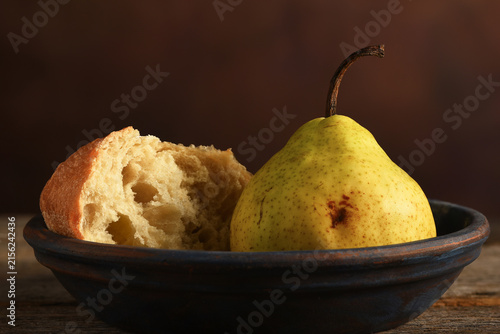 Pear and Bread Still Life. An Anjou Pear and a piece of broken bread on a plate and rustic wooden table with a mottled brown background.