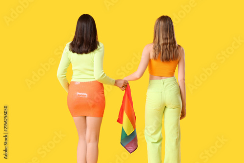 Young lesbian couple with LGBT flag on yellow background, back view