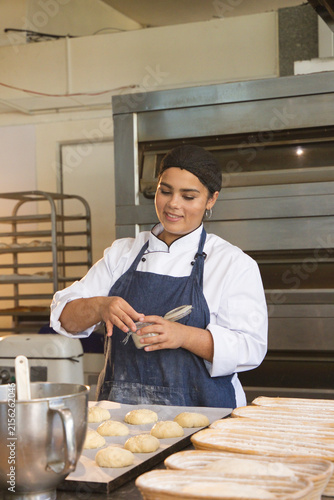 Fotografía African American female baker brushing dough in coat at counter with trays proof